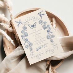 a white and blue wedding card sitting on top of a plate next to some feathers