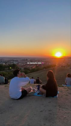 two people sitting on the ground at sunset eating and watching something in front of them