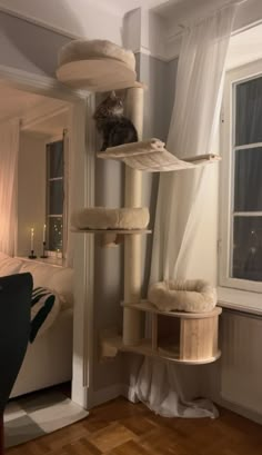 a cat sitting on top of a scratching tower in a bedroom next to a window