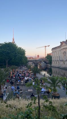 many people are sitting on benches near the water and some buildings with cranes in the background