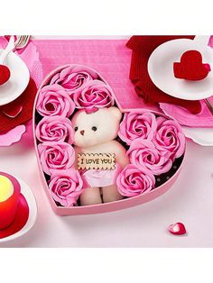 a teddy bear in a heart shaped box surrounded by pink roses and candles on a table