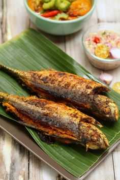 grilled fish on a banana leaf next to bowls of vegetables