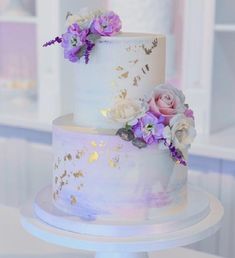 a three tiered white cake with purple and white flowers on the top, sitting on a table