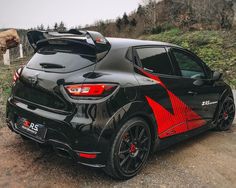 the rear end of a black and red sports car parked on a dirt road with trees in the background