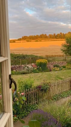 an open window looking out onto a garden and field in the distance with trees, shrubs, and flowers