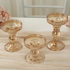 three glass candlesticks sitting on top of a white table cloth next to pink flowers