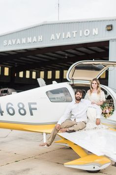 a man and woman sitting on the wing of a small plane in front of a hangar