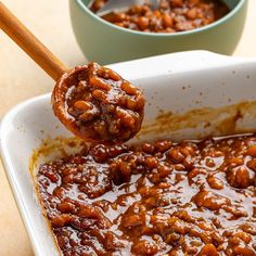 a spoonful of baked beans is being lifted from a casserole dish with another bowl in the background