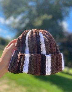 a hand holding up a brown and white striped knitted beanie in front of a tree