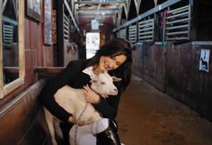 a woman holding a lamb in her arms while standing next to a horse stall door