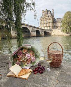 a bouquet of flowers sitting on the ground next to a wine glass and book near a river with a bridge in the background