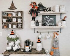 An off-white wall features light wood shelving containing Rae Dunn jars and halloween decor. Beside the shelves hangs an antique apothecary display unit with halloween decor and trinkets. Below, a coffee station sits on a wooden tea trolley including a coffee machine, mug tree and faux foliage...   Image: decor_doctor