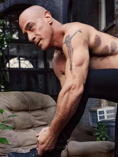 a shirtless man with tattoos on his arm and leg, leaning over a couch