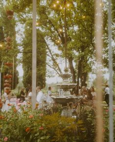 people are gathered around a fountain in the middle of a garden with flowers and trees