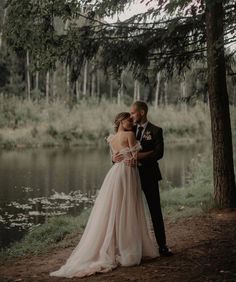 a bride and groom standing next to each other in front of a lake surrounded by trees