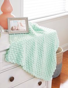 a baby blanket on top of a white dresser next to a pink vase and photo frame