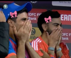 two men covering their faces with pink bows on their heads at a sporting event in india