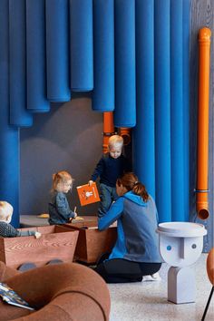 three children are sitting at a table in front of blue walls and chairs, while one child is holding an orange book