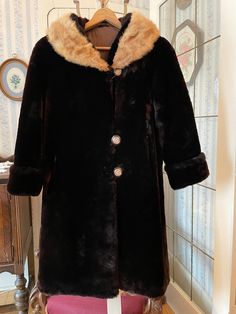 "This gorgeous, heavy coat is made from super warm mouton in very dark rich brown, complemented by a light golden brown mink fur collar. It fastens with the original decorative buttons in the front and has two front pockets and one inside pocket with the initials \"MER.\" It is fully lined with brown satiny lining. The measurements, taken with the coat lying flat, are: shoulder to shoulder, 17inches; armpit to armpit, 19 inches; sleeves, 22 inches; length, 40 inches; bottom edge, 32 inches. In v Elegant Brown Long Fur Coat, Elegant Long Brown Fur Coat, Brown Winter Fur Coat For Formal Occasions, Brown Fur Coat For Formal Winter Occasions, Classic Brown Fur Coat With Faux Fur Trim, Classic Brown Long Fur Coat, Classic Long Brown Fur Coat, Brown Long Coat With Faux Fur Trim, Vintage Fur Coat