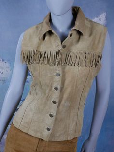 This tan leather vest has a wing collar, and closes in the front with seven bronze buttons. The straight-front Western style cowboys and Indians vest has a row of 2.5-inch (6.35cm) fringe across the bust line, and features corset-style lacing on each side. Bust = 38 inches (96.52cm) Vest Length = 21.5 inches (54.61cm) Size: 10 US, 14 UK Material: Genuine Leather --- CONDITION --- This tan leather vest is vintage, but is in Excellent condition, and is clean, soft, and supple. There are no tears, Western Leather Vest For Fall, Brown Sleeveless Vest For Western-themed Events, Fitted Vest For Western-themed Events In Fall, Western Style Brown Vest For Western-themed Events, Brown Western Vest For Western-themed Events, Fitted Western Leather Vest, Fitted Western Vest For Spring, Fitted Leather Festival Vest, Western Style Fitted Vest For Spring