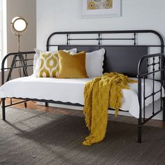 a metal day bed with yellow pillows and blankets on it's sides, against a white brick wall