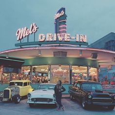 a woman standing in front of a drive - in with old cars parked outside it