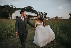 a bride and groom holding hands walking through tall grass in front of a farm house