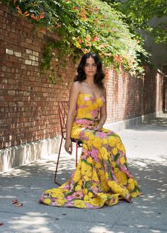 a woman is sitting on a chair in front of a brick wall wearing a yellow floral dress