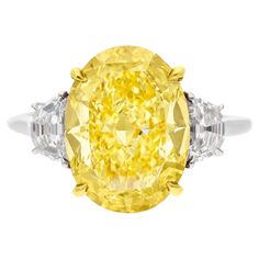 This exquisite ring features a captivating GIA certified 6.01 carat Fancy Yellow oval cut diamond as its centerpiece. The diamond’s Fancy Yellow color grade showcases a delicate and charming hue, adding a touch of warmth and uniqueness. With a clarity grade of VS1, the diamond has very slight inclusions that are not visible to the naked eye, ensuring exceptional clarity. The diamond’s polish is graded as Excellent, providing a flawlessly smooth surface that maximizes its brilliance. Its symmetry is rated as Very Good, indicating precise and well-balanced faceting that enhances the diamond’s overall sparkle. Additionally, the diamond exhibits faint fluorescence, allowing its natural color and clarity to shine without any interference. Flanking the central oval diamond are two beautifully cu Angie Crabtree, Fancy Yellow Diamond Ring, Radiant Diamond Rings, Radiant Cut Diamond Ring, Canary Diamond, Yellow Diamond Ring, Cushion Diamond Ring, Yellow Diamonds Engagement, Cushion Cut Diamond Ring