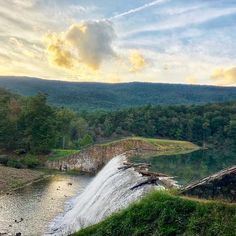 a large waterfall in the middle of a river surrounded by trees and hills under a cloudy sky