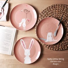 three pink plates with rabbits on them and an open book next to each other, sitting on a wooden table