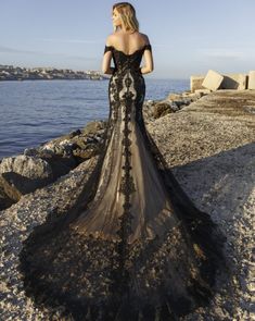 a woman in a black dress standing on the rocks by the water with her back to the camera