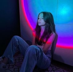 a woman sitting on the floor in front of a wall with colorful lights behind her