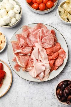 several different types of meats and vegetables on a white plate with bowls of olives