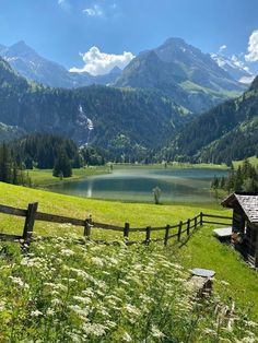 a wooden fence is in front of a mountain lake and grassy field with wildflowers