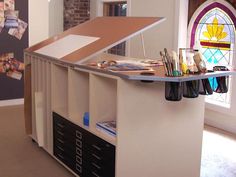 an office desk with lots of art supplies on the top and bottom shelf, in front of a stained glass window