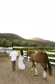 a man and woman walking with a horse on a dirt road in front of a white fence