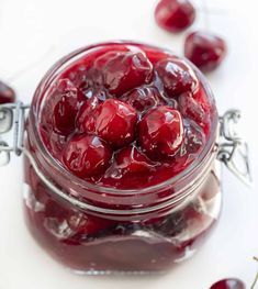 a glass jar filled with cherries on top of a white table next to green leaves