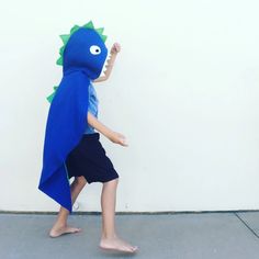 a young boy wearing a blue dinosaur cape and black shorts, standing in front of a white wall