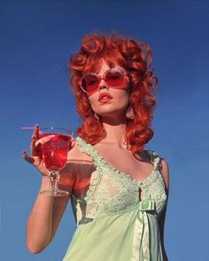 a woman with red hair wearing sunglasses and holding a drink in her hand, against a blue sky