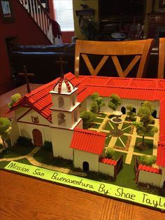 a model of a house with a red roof and white trim on the top, sitting on a table in front of a wooden chair