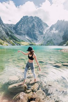 a woman standing on rocks in the middle of a lake with mountains in the background