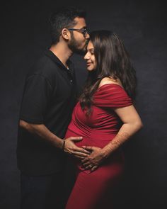 a pregnant woman and man pose for a photo in front of a dark background with their arms around each other