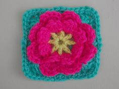 a crocheted square with a pink flower on the center is hanging from a hook