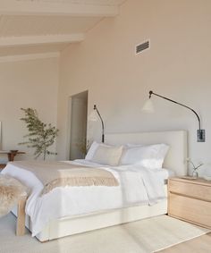 a bed with white sheets and pillows in a bedroom next to a dresser, lamp and window