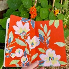 a hand holding an open notebook with flowers on it in front of some green leaves