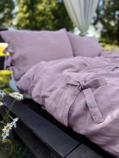 an unmade bed with purple linens and daisies in the foreground, outside