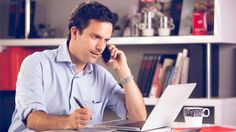 a man sitting in front of a laptop computer talking on a cell phone and holding a pen