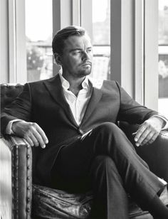 a black and white photo of a man in a suit sitting on a couch looking out the window