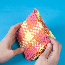 two hands are holding an orange and pink woven object on a blue surface with one hand reaching for it